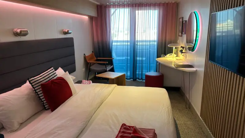 Virgin Voyages cruise ship cabin with a modern design, featuring a bed with red and striped pillows, ambient lighting, a chair, a desk with a lamp, a circular mirror, and a balcony with sheer curtains.