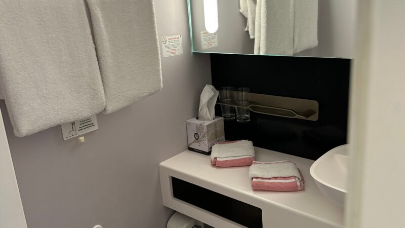 Virgin Voyages cruise ship cabin bathroom with a white toilet, sink, illuminated mirror, and towels neatly arranged.