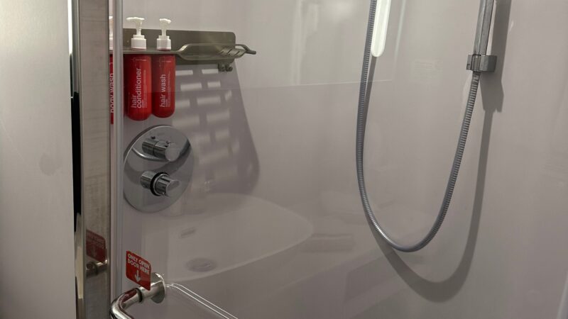 Shower area in a Virgin Voyages cruise ship cabin with a glass door, chrome fixtures, and red shampoo and conditioner bottles mounted on the wall.