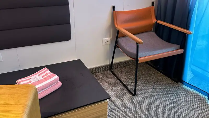 Cruise ship cabin interior featuring a chair, a bed with folded towels, a nightstand, and curtains.