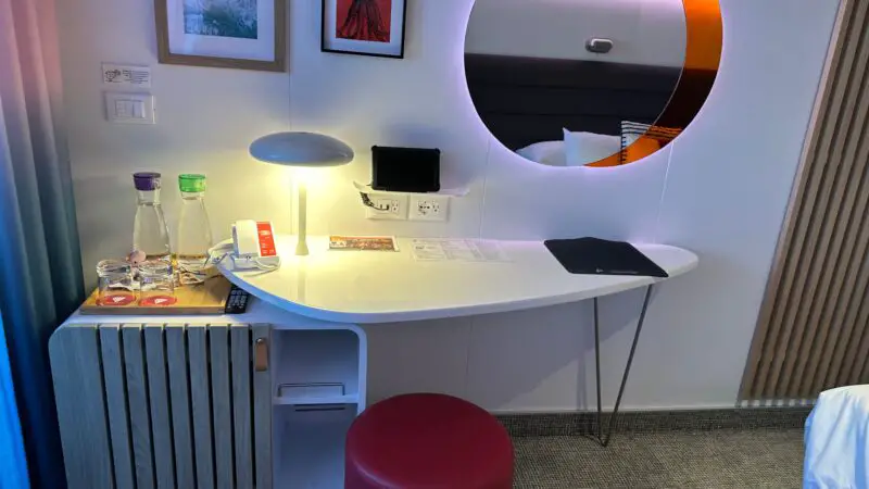 Virgin Voyages cruise ship cabin with a desk, two water carafes, a lamp, framed art, and a round mirror, next to a bed.