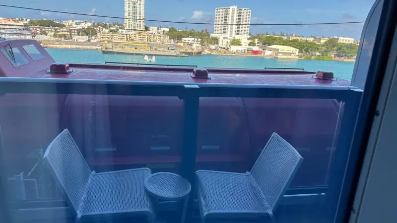 Virgin Voyages cruise ship balcony cabin view, showing two chairs and a table with a coastal cityscape in the background.