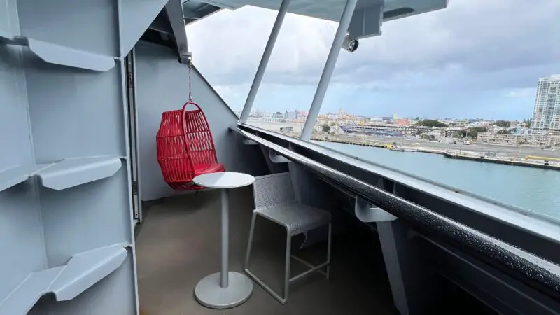 Virgin Voyages cruise ship balcony with a red hanging chair, two gray chairs, and a small round table, overlooking a port.