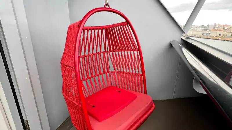 Cruise ship balcony featuring a red hanging chair with cushions, overlooking a cityscape.