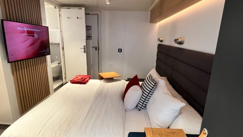Virgin Voyages cruise ship cabin with a bed, pillows, and nightstands, featuring modern decor and a wall-mounted TV.