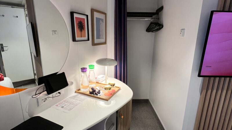 Virgin Voyages cruise ship cabin featuring a compact desk with a lamp, water bottles, and snacks on a tray. A flat-screen TV is mounted on the wall above a vertical wooden panel. A closet with hangers and framed art are visible.