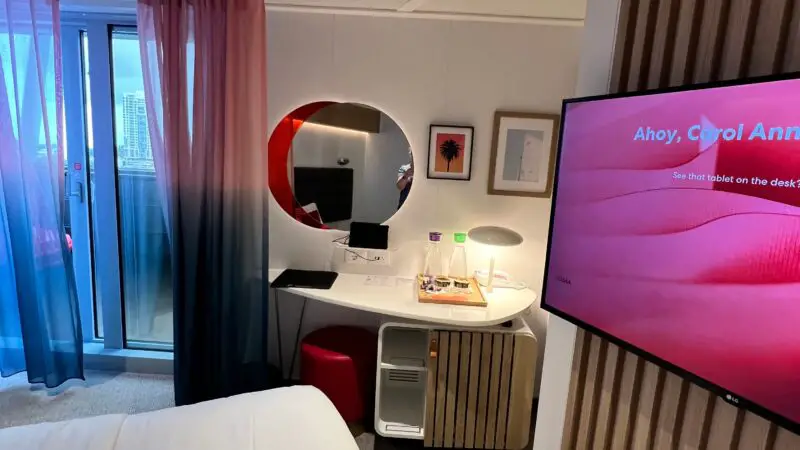 Interior of a Virgin Voyages cruise ship cabin showing a bed, wall-mounted TV, desk with a mirror, and balcony with sheer curtains.