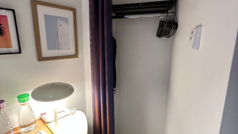 Virgin Voyages cruise ship cabin with a small desk, lamp, bottled water, and an open closet with hangers, framed art on the wall.