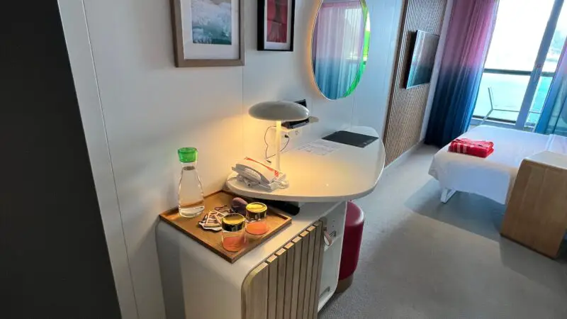 Cruise ship cabin interior with a desk, lamp, wall art, round mirror, and balcony view.