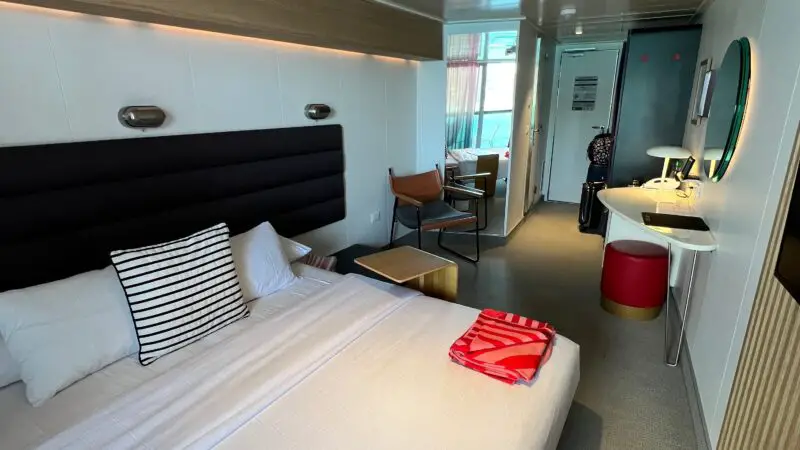 Virgin Voyages cruise ship cabin featuring a bed with striped pillows, a small table and chairs, a mirror above a desk, and a glimpse of a balcony with curtains.