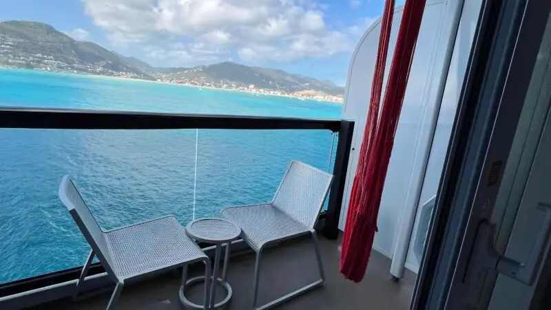View from a Virgin Voyages cruise ship balcony showing two chairs and a small table, overlooking the ocean and distant mountains.