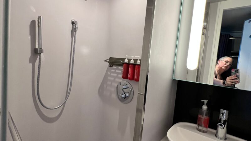 A Virgin Voyages cruise ship stateroom bathroom showing a shower with a rainfall showerhead, handheld shower wand, toiletries on a shelf, and a sink with a mirror.