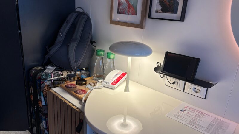 Cruise cabin interior with a sleek desk, phone, lamp, tablet on a wall mount, framed art, bottles, and luggage.