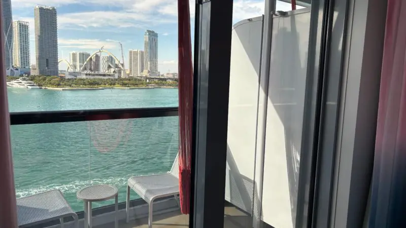 View from a Virgin Voyages cruise ship cabin showing a private balcony with two chairs and a small table overlooking the water and cityscape.