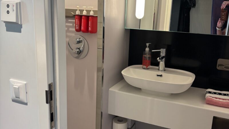Bathroom of a Virgin Voyages cruise ship cabin featuring a modern sink, mirror, and shower area with red toiletries.