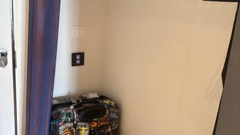 Cruise ship cabin closet with a suitcase, backpack, hangers, and shelving on Virgin Voyages.