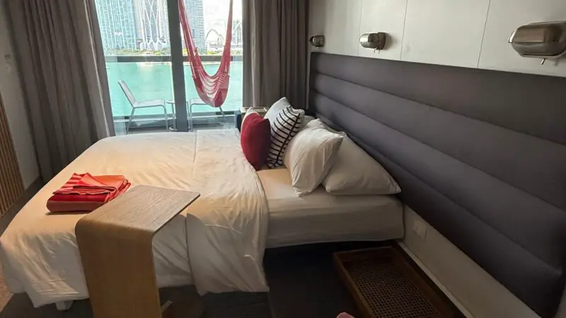 Virgin Voyages cruise ship cabin featuring a bed, striped pillows, red hammock outside on the balcony, and a view of the ocean and city skyline through sliding glass doors.