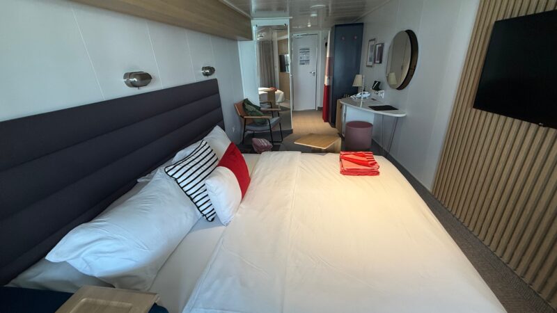 Cruise ship cabin with a bed, striped cushions, wall-mounted TV, round mirror, and a small table with chair.