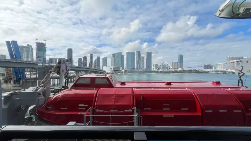 View from a Virgin Voyages cruise ship balcony, overlooking a red lifeboat and a city skyline with tall buildings under a partly cloudy sky.