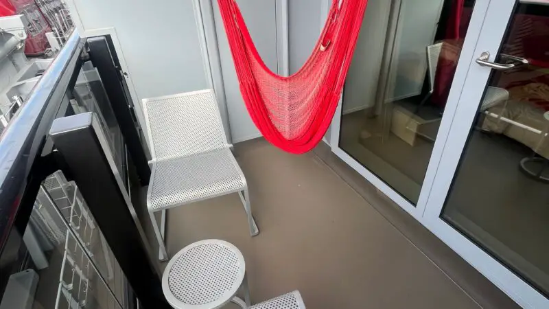Cruise ship cabin balcony with two white chairs, small round table, and a red hammock.