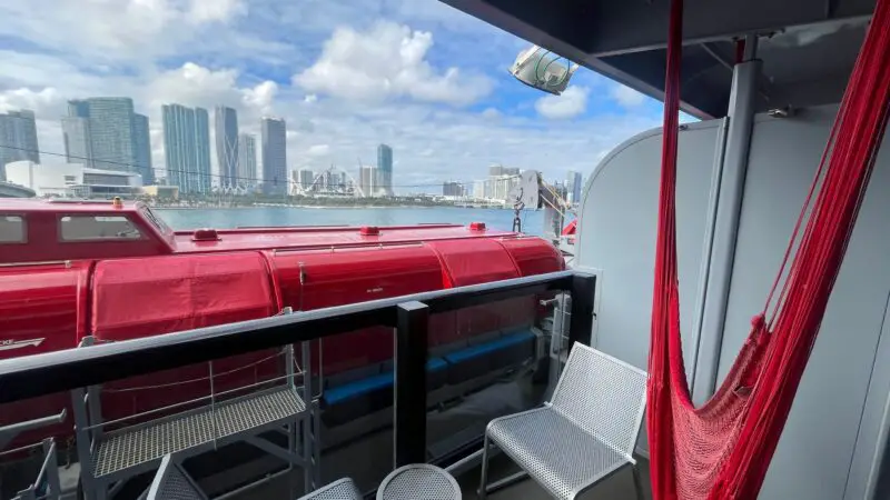 Virgin Voyages cruise ship balcony with a red hammock, chairs, a small table, and a view of a city skyline across the water.