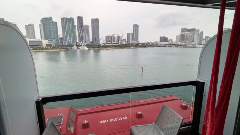Cruise ship balcony with two chairs, a small table, and a hammock, overlooking a city skyline across the water.