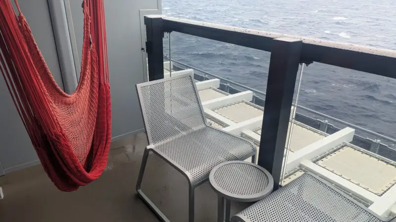Cruise ship balcony with a red hammock, two metal chairs, and a small table overlooking the ocean.