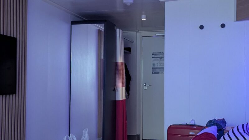 Virgin Voyages cruise ship cabin with a bed, wooden bedside table, red and striped pillows, and a suitcase near the door.