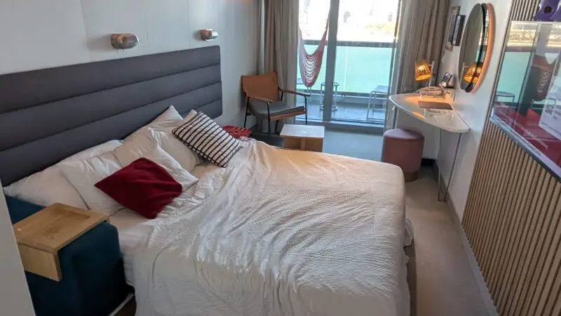 Cruise ship stateroom with a large bed, wall-mounted TV, seating area, and a balcony with a hammock overlooking the water.