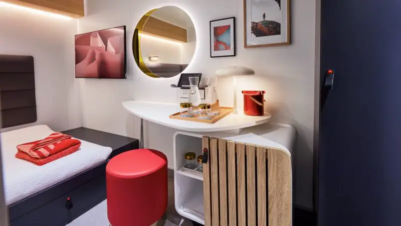 Modern cruise cabin with circular backlit mirror, desk, and red ottoman on a Virgin Voyages cruise ship