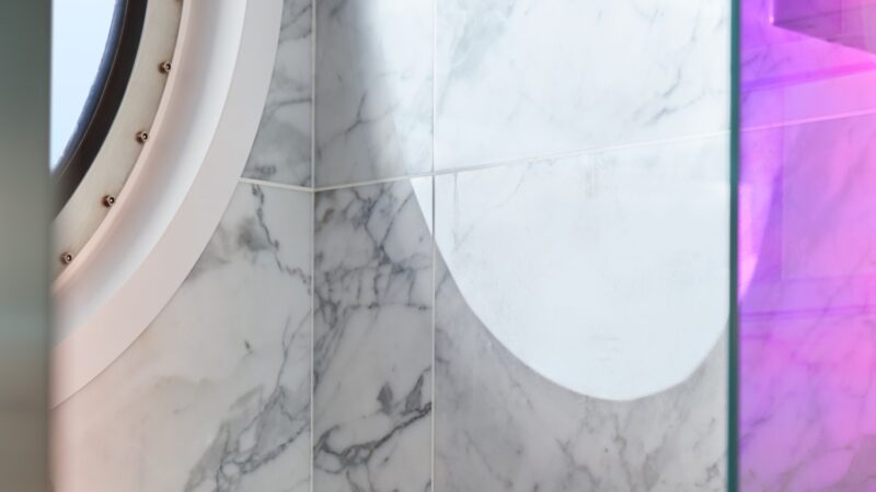 Circular porthole window in a marble-tiled cabin wall on a Virgin Voyages cruise ship with purple lighting