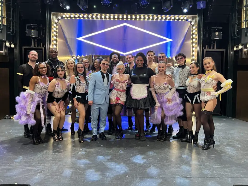 Group of performers in colorful show costumes posing on stage with illuminated diamond backdrop