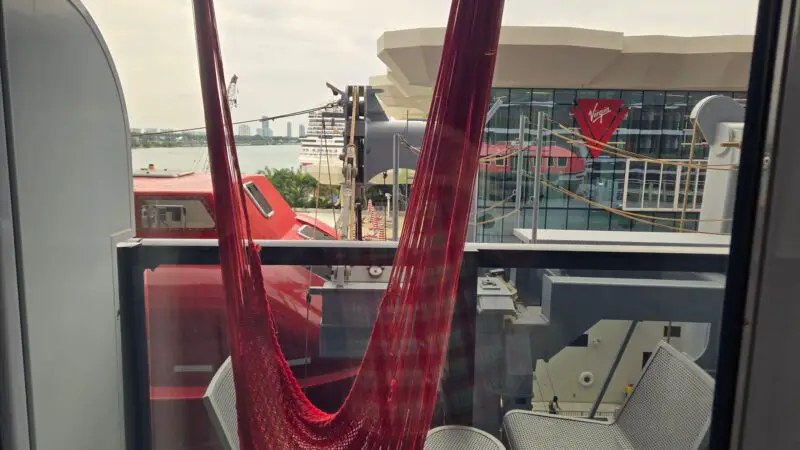View from a Virgin Voyages cruise ship balcony cabin, showing a red hammock and chairs on the balcony, with the Virgin terminal and water in the background.