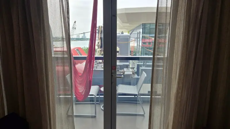 Virgin Voyages cruise ship cabin with glass doors leading to a balcony; a red hammock and chairs are visible outside.