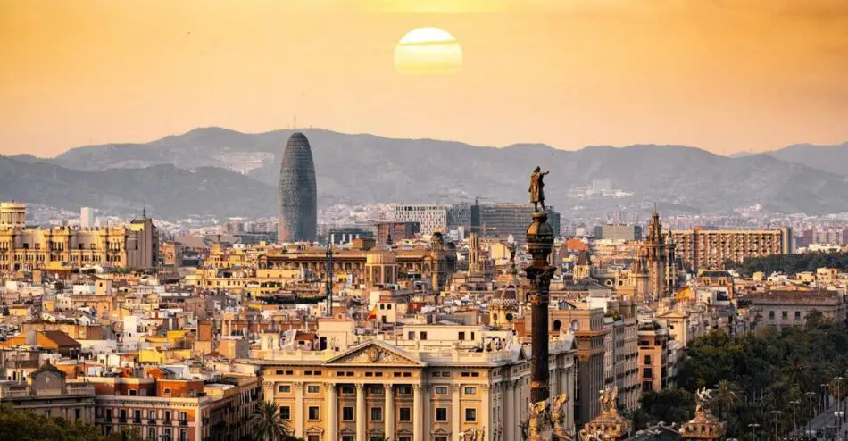 Cityscape with a prominent tall statue, modern skyscraper, and mountains in the background against an orange sunset.
