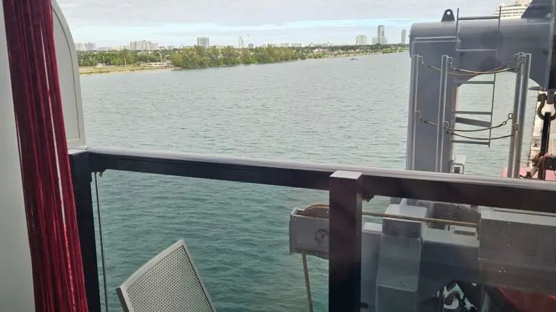 A Virgin Voyages cruise ship balcony cabin with two metal chairs, a small table, and a view of the sea and distant shoreline.