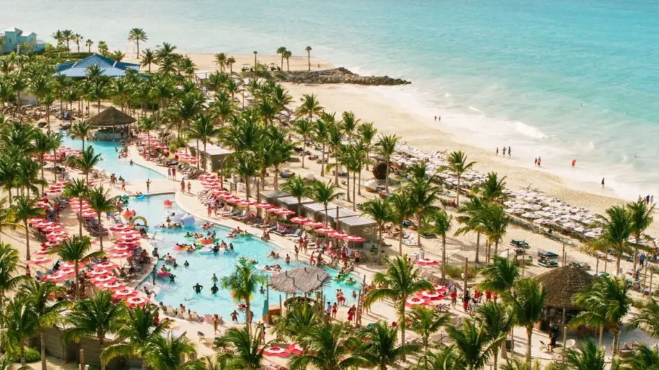 Aerial view of a beach resort with a pool, red umbrellas, and palm trees by the ocean.