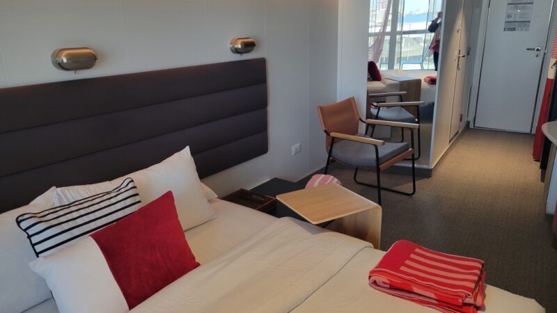 Virgin Voyages cruise ship cabin with a neatly made bed, striped and red pillows, a small table, and a chair. A balcony with curtains is visible in the background.