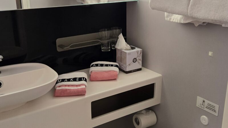 Cruise ship cabin bathroom with a modern sink, mirror with light, folded towels, box of tissues, and a toilet.