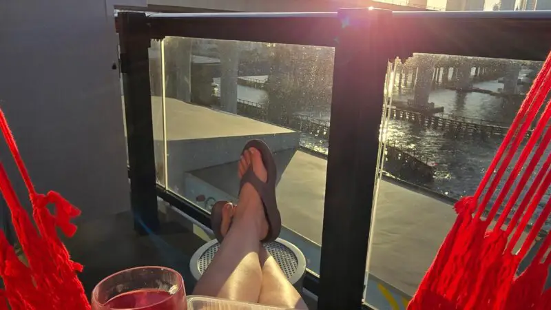 Person relaxing on a Virgin Voyages cruise ship balcony hammock with snacks and a drink, overlooking a sunset-lit cityscape.