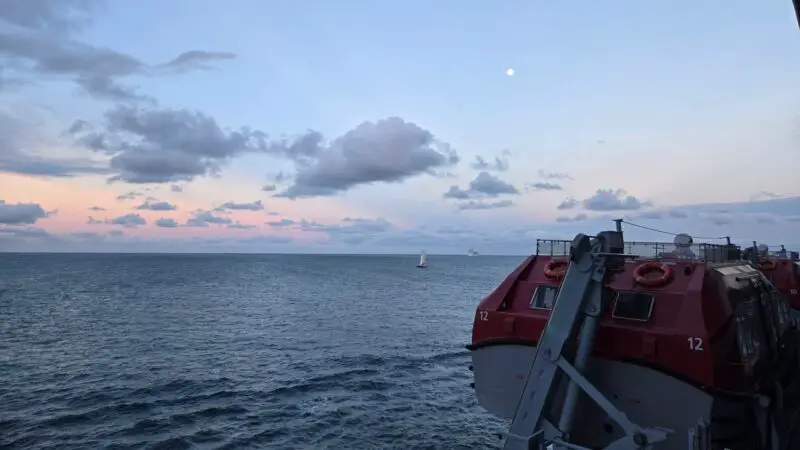 View from a Virgin Voyages cruise ship balcony, showing the open ocean at sunset with a lifeboat in the foreground and a sailboat in the distance.