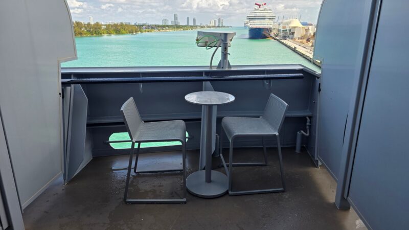 Balcony view from a Virgin Voyages cruise ship cabin showing two chairs and a small table, overlooking turquoise water and a distant ship at port.