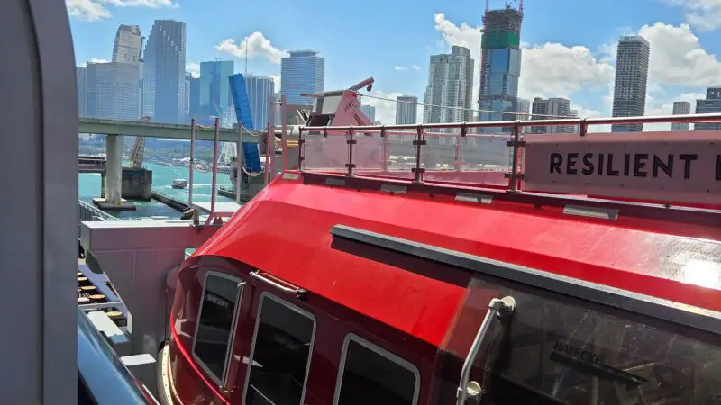 View from a Virgin Voyages cruise ship balcony showing a red lifeboat and a city skyline under a blue sky.