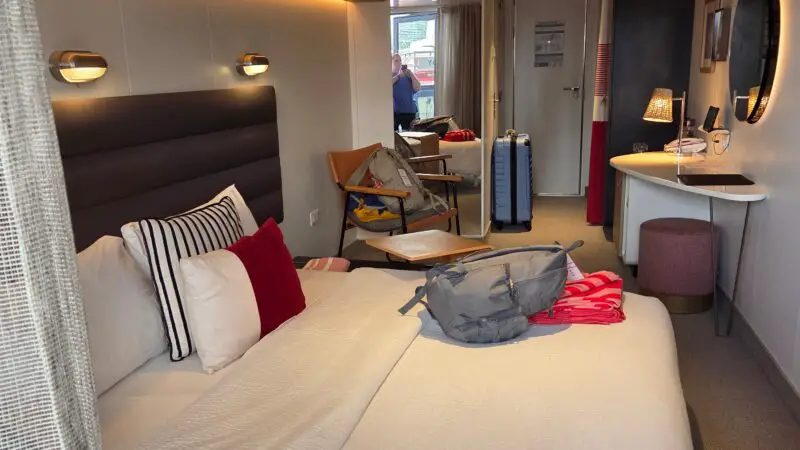 Interior view of a Virgin Voyages cruise ship cabin with a neatly made bed, two-tone pillows, a small desk with a round mirror, and a sliding door leading to a balcony.