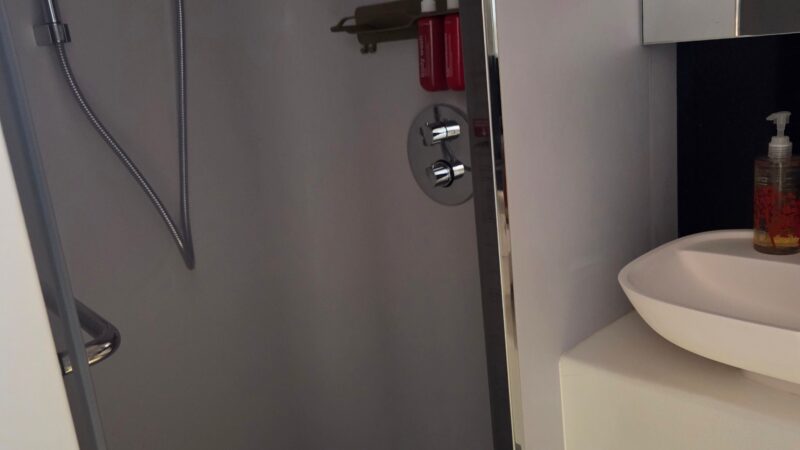 Shower area in a Virgin Voyages cruise ship cabin bathroom, featuring a hand-held showerhead and shelf with toiletries.