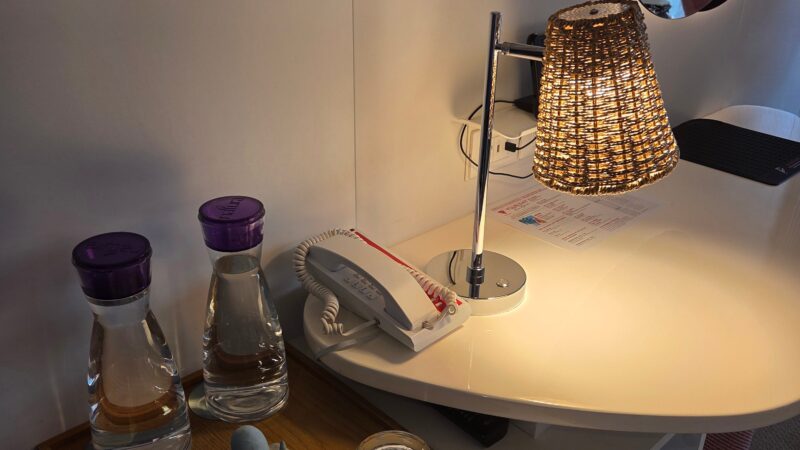 Virgin Voyages cruise ship cabin showing a desk with a decorative lamp, phone, water bottles, glasses on a tray, and framed artwork on the wall.