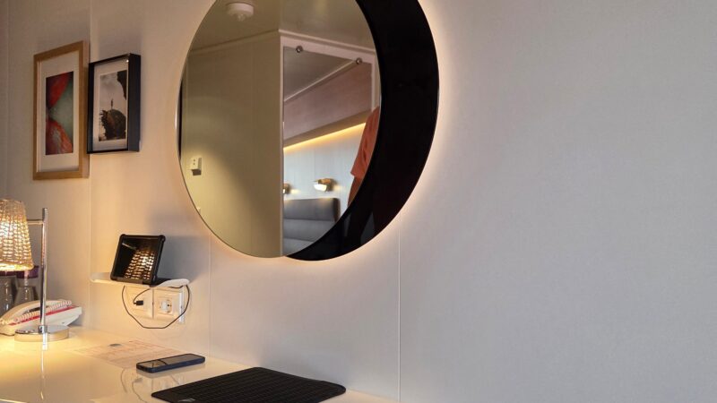 Virgin Voyages cruise ship cabin interior with a modern desk, round mirror, wall art, and upholstered stool.