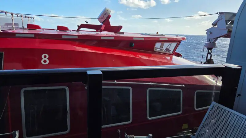 View from Virgin Voyages cruise cabin balcony showing ocean and red ship structure with numbered section. A chair and mug are visible in the foreground.