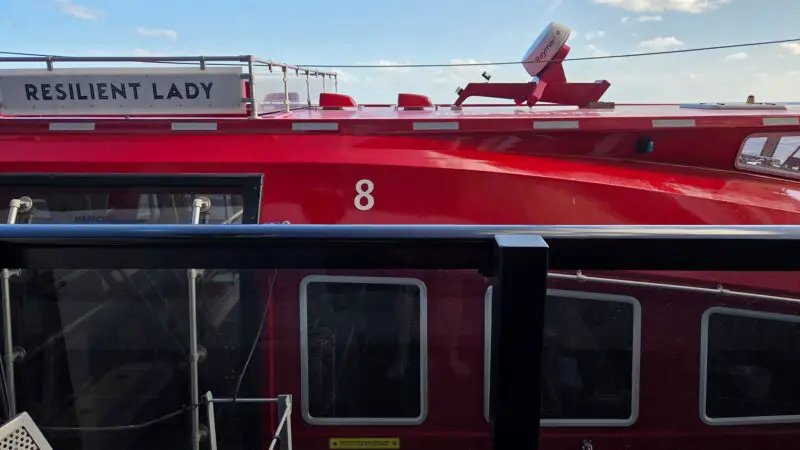 View from a Virgin Voyages cruise ship balcony, showing the red exterior with “Resilient Lady” and number “8” visible, under a partly cloudy sky.