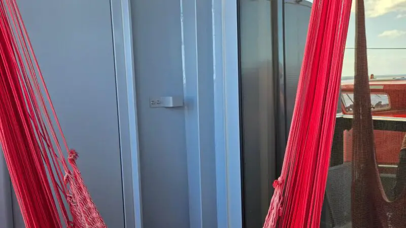 A Virgin Voyages cruise ship cabin balcony with a red hammock hanging beside a sliding glass door, showing a view of the sky with some clouds.